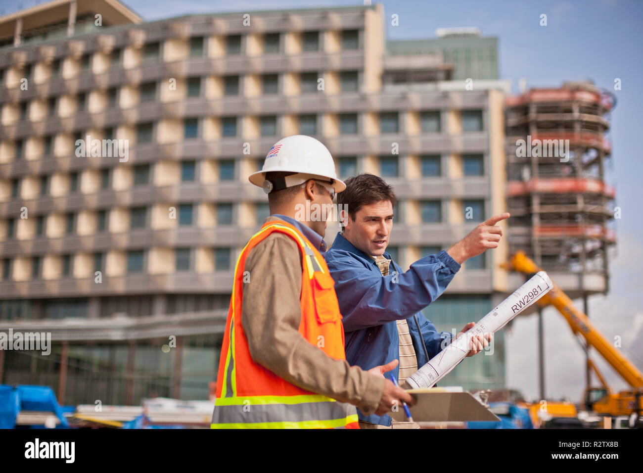 Two engineers in high visibility vests and hardhats discuss building plans on a construction site. Stock Photo