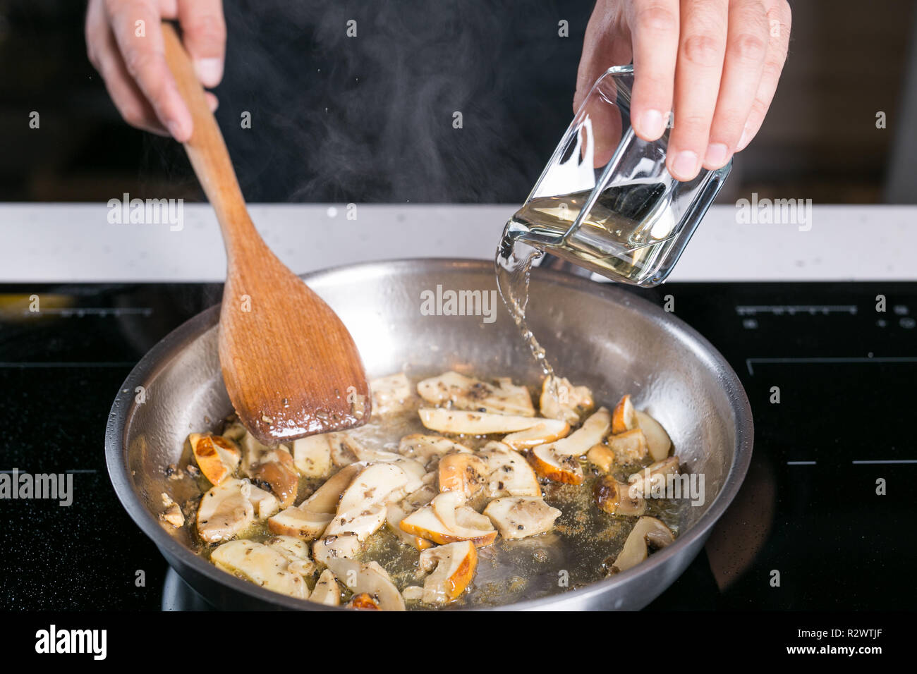 Chef pouring transparent liquid over cooking mushrooms in a metal pan