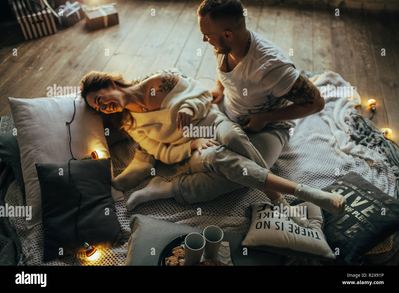 Young couple in love having fun and laughing cheerfully on background of wooden floor, coverlet, pillows and glowing lightbulbs. Stock Photo