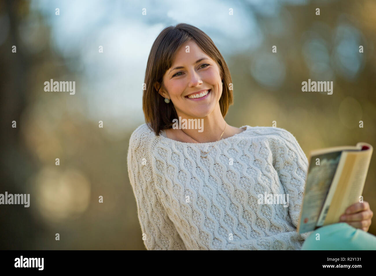 Smiling young woman relaxing with a book in a sunlit garden. Stock Photo