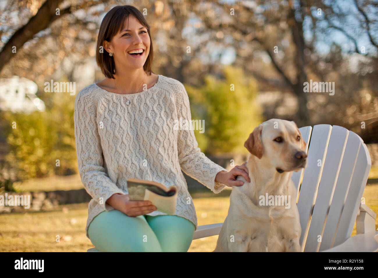 Smiling young woman relaxing with her dog and a book in a sunlit garden. Stock Photo