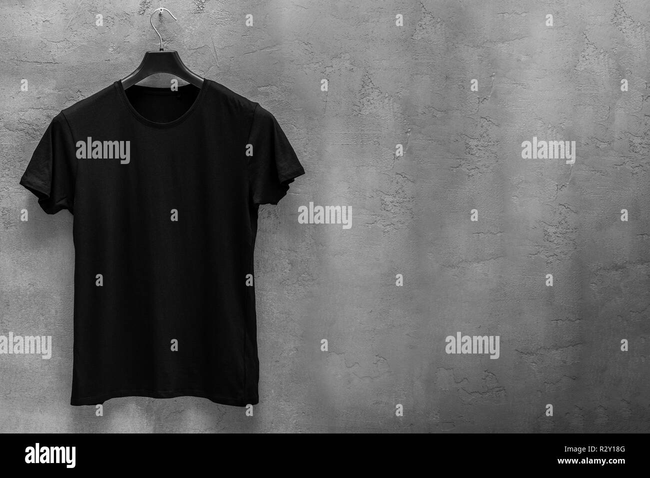 Front side of male black cotton t-shirt on a hanger and a concrete wall in the background. T-shirt without print and copyspace for your text on right Stock Photo