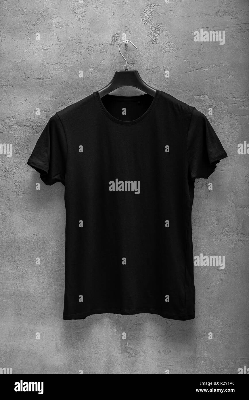 Front side of male black cotton t-shirt on a hanger and a concrete wall in the background. T-shirt without print Stock Photo
