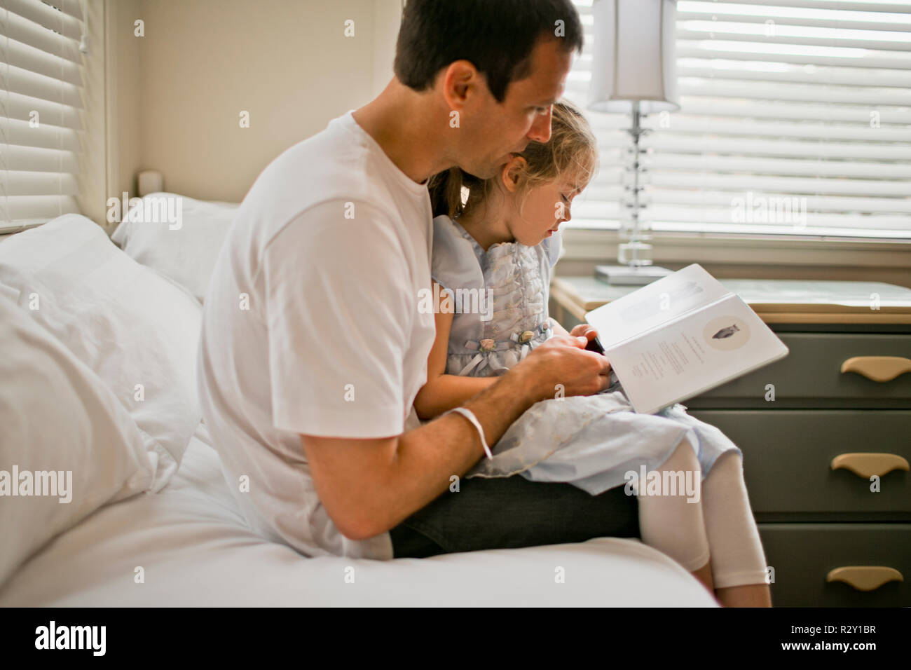 Father sitting on a bed reading a book with his daughter. Stock Photo
