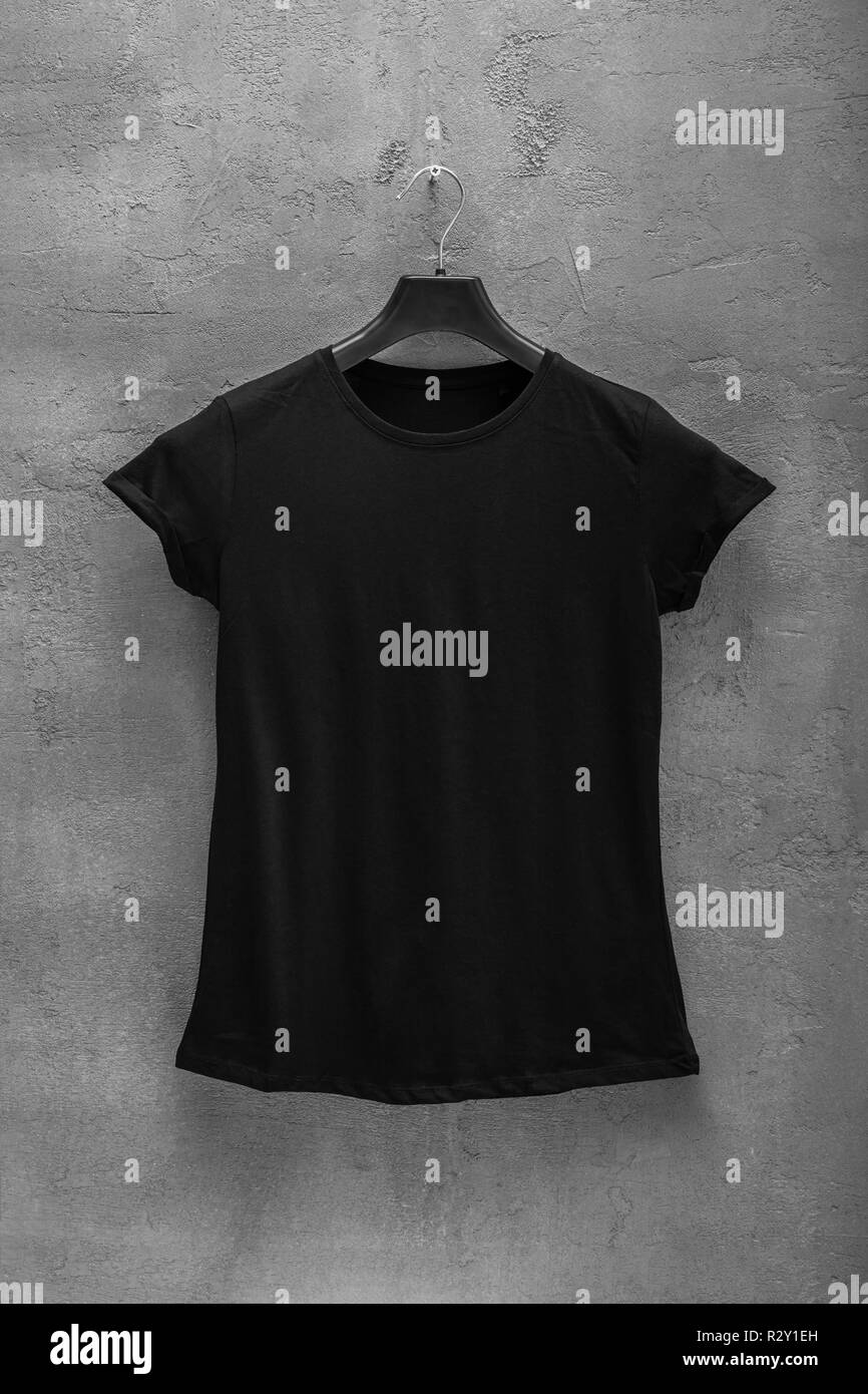 Front side of female black cotton t-shirt on a hanger and a concrete wall in the background. T-shirt without print Stock Photo