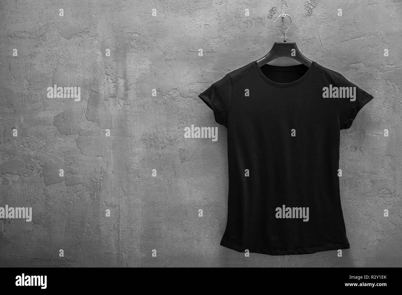 Front side of female black cotton t-shirt on a hanger and a concrete wall in the background. T-shirt without print and copyspace for your text on left Stock Photo