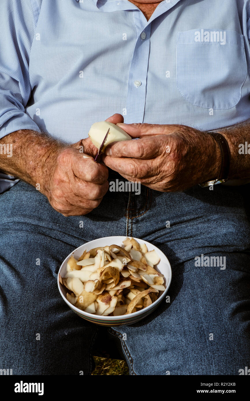 A man seated using a sharp knife to peel potatoes. Stock Photo