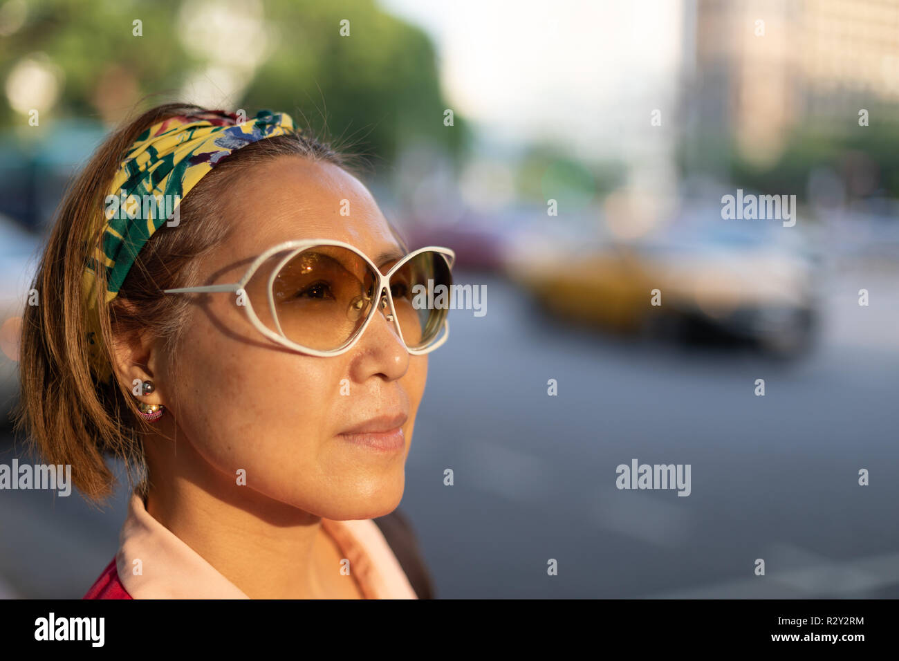 Portrait of confident mature Taiwanese woman of Chinese ethnicity looking into the distance in the evening sun against big city traffic backdrop Stock Photo