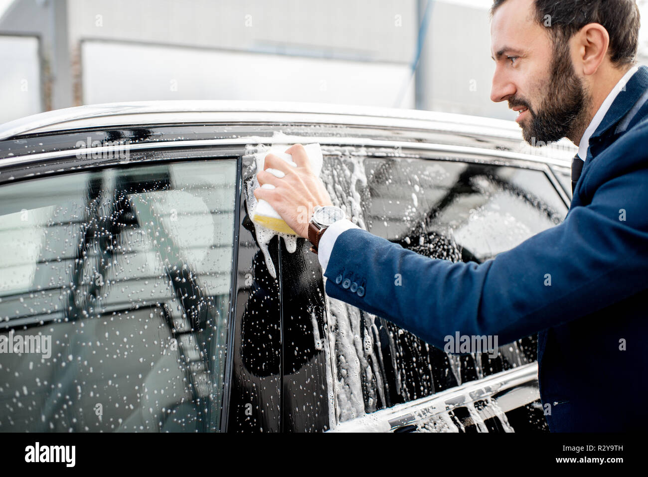 Elegant businessman dressed in the suit washing his car with yellow sponge on a self service open air car wash Stock Photo