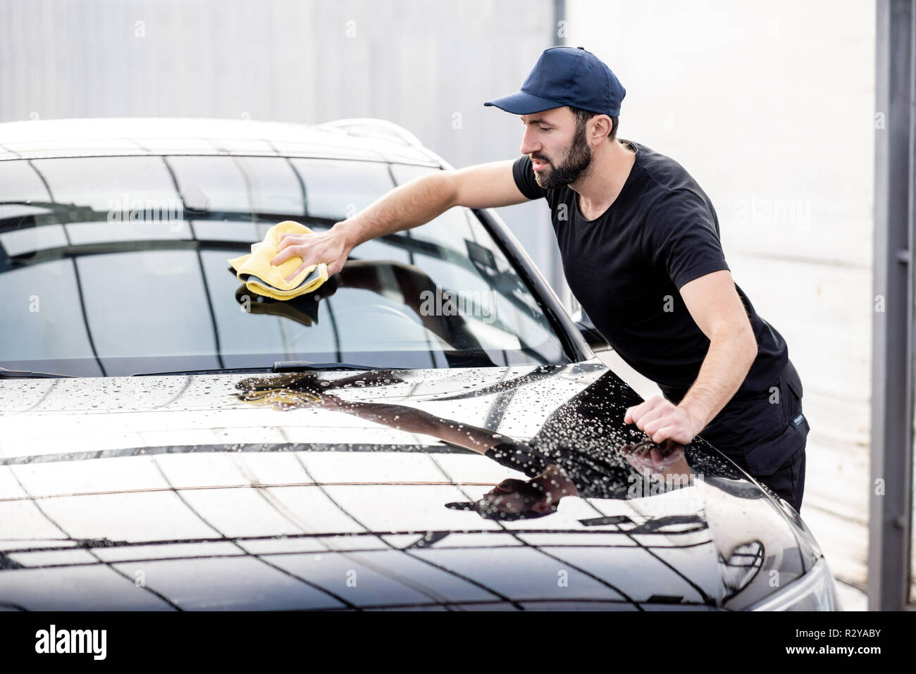 Professional washer in t-shirt and cap wiping windshield with yellow microfiber at the open air car wash Stock Photo