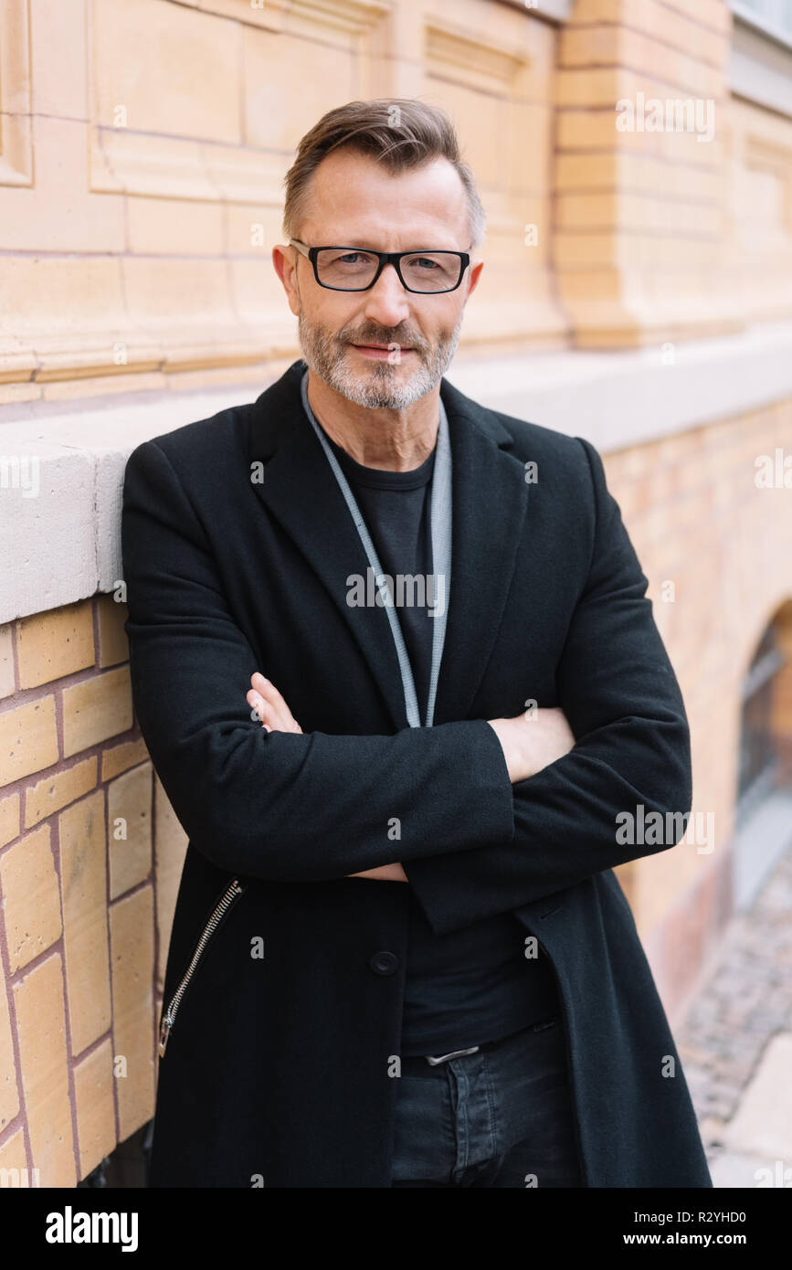 Serious confident middle-aged man with folded arms standing next to an urban brick wall staring intently at the camera Stock Photo
