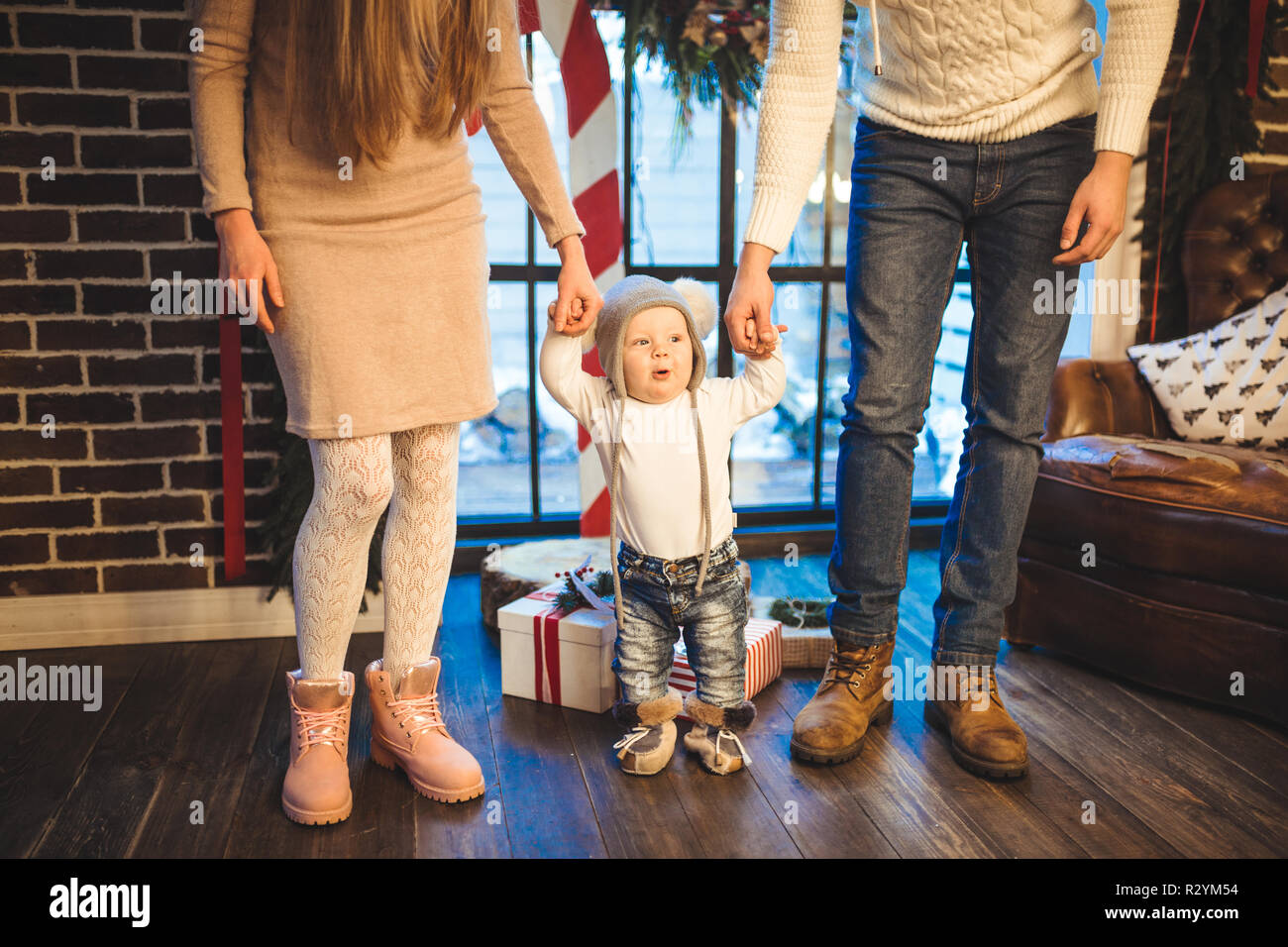 Funny little baby boy 1 year old learning walk home in winter in a decorated New Year house. Young family dad and mom hold by the hands of his son in  Stock Photo
