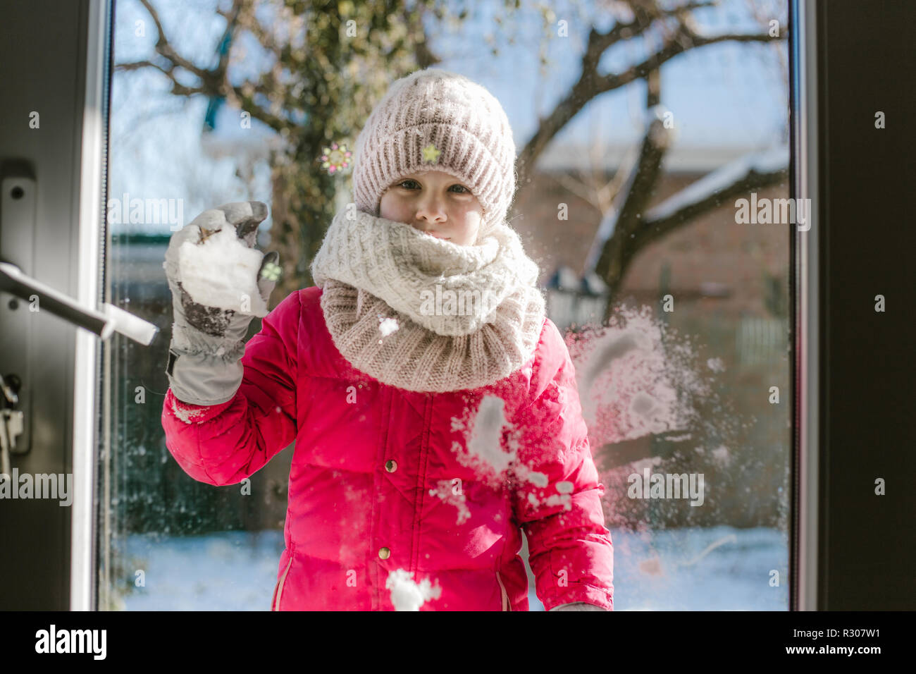 Seven-year-old cute girl in winter clothes is standing outside the door, on the street with snow in her hands and looking into the house smiling. Stock Photo