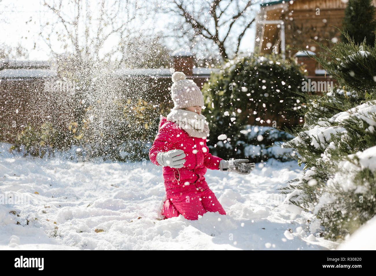 Seven-year cute girl in winter clothes playing with snow in the backyard of a house on a winter sunny day Stock Photo