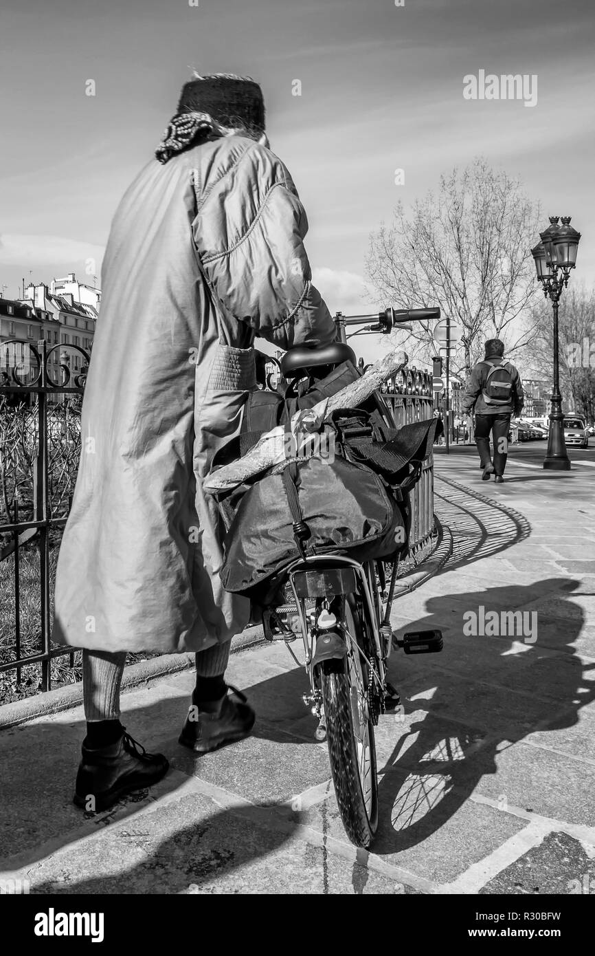 Black and white view of a woman with a bicycle and a typical French baguette on the streets of central Paris, France Stock Photo