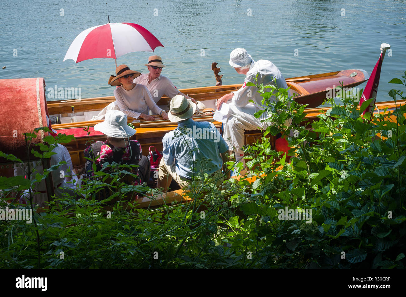 A red and white umbrella shelters a party on two classic vintage mahogany boats by the towpath at Henley Royal Regatta, Henley-on-Thames, Oxfordshire Stock Photo