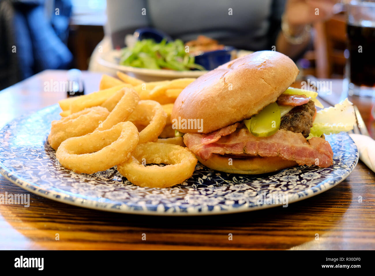 A JD Wetherspoons Ultimate Burger meal served with onion rings and chips. The meal is on a pub table in a busy Wetherspoons pub Stock Photo