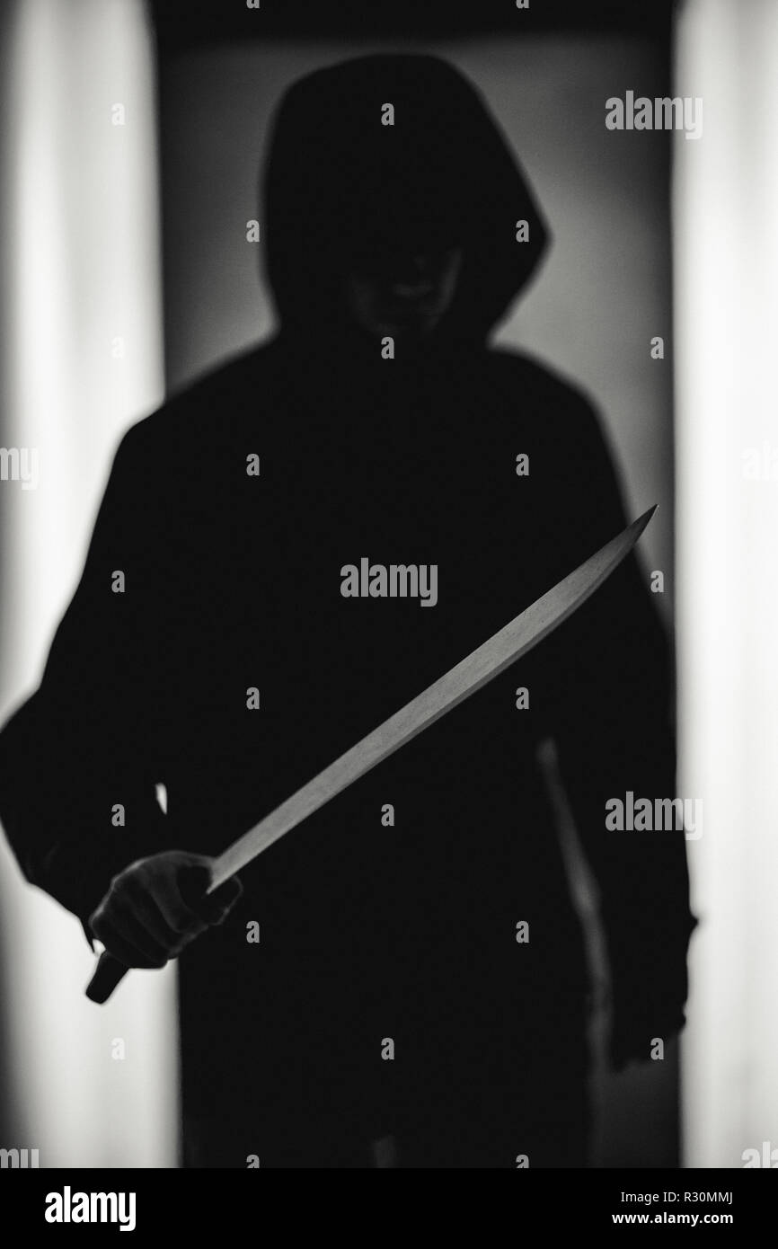 Black and white photo of criminal in hood with machete in dark room Stock Photo