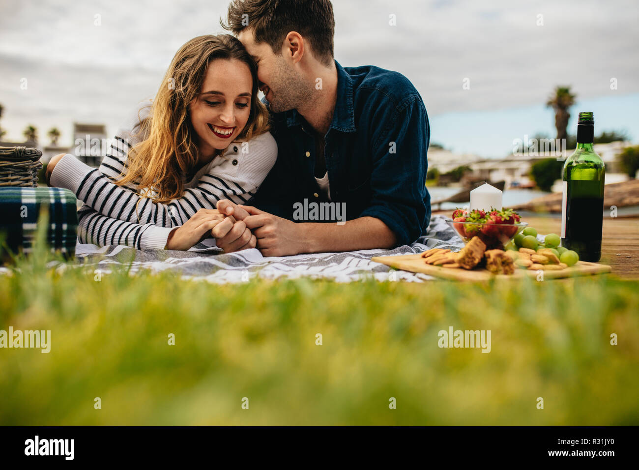 Couple in love on a date enjoying food and drinks sitting outdoors. Smiling man and woman lying down on the ground holding hands and talking while on Stock Photo