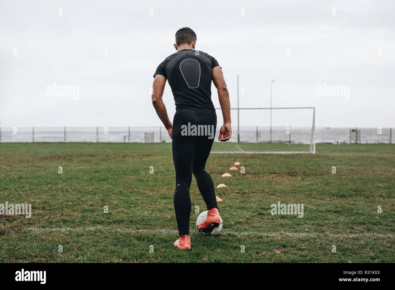Rear view of a soccer player practicing dribbling with the help of cones arranged on field. Football player practicing on field early in the morning. Stock Photo