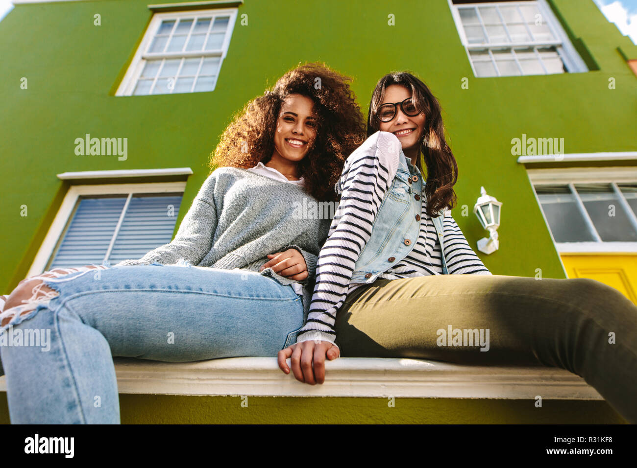 Low angle view of two women sitting on bench outside a green house. Female friends hanging out outdoors looking at camera. Stock Photo