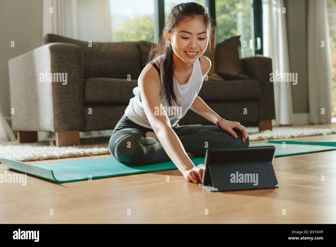 Smiling woman sitting exercise mat and watching training videos on digital tablet. Young chinese female in sportswear exercising watching online video Stock Photo