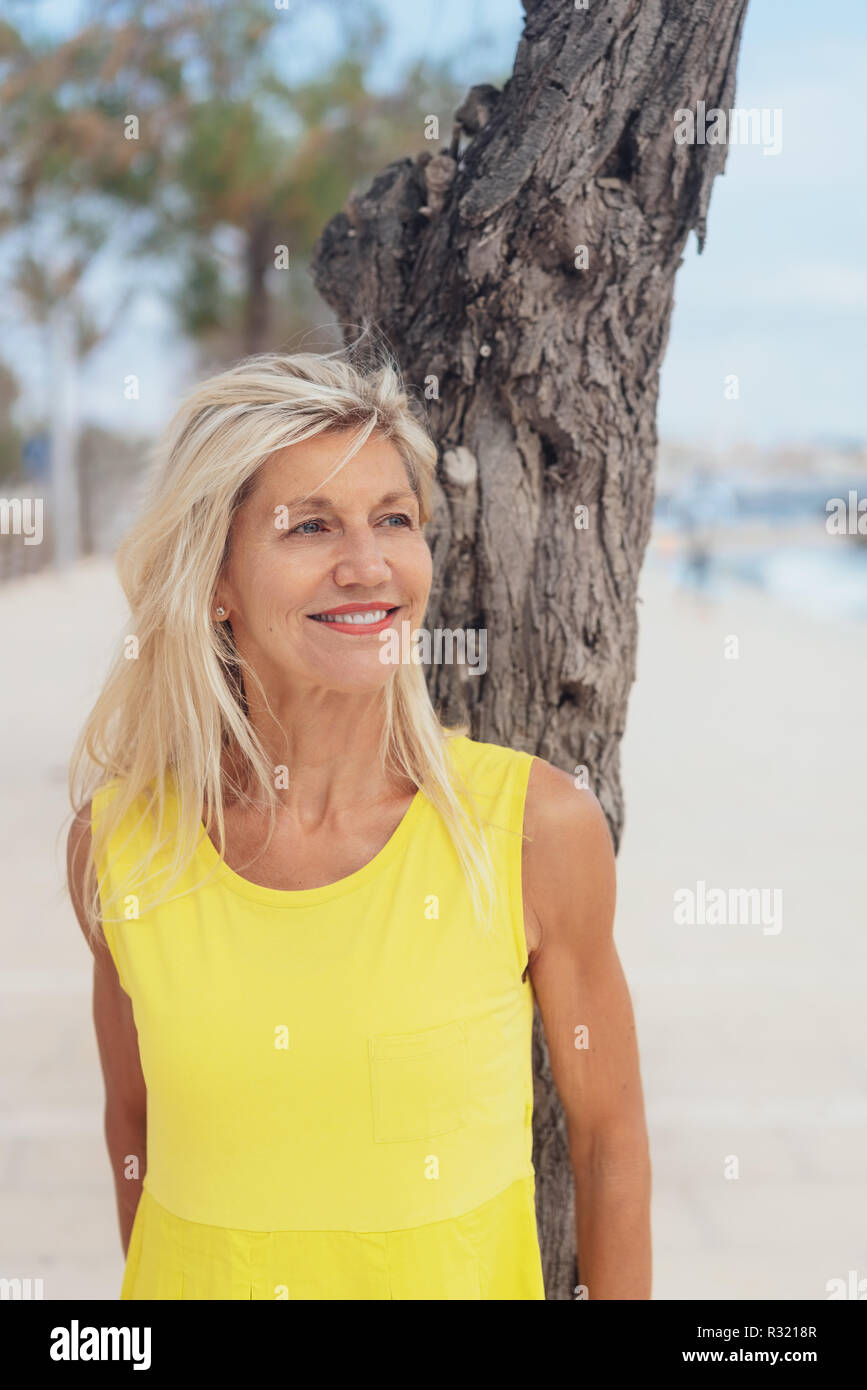 Attractive older woman standing leaning on a tree trunk at the beach looking to the side with a smile Stock Photo