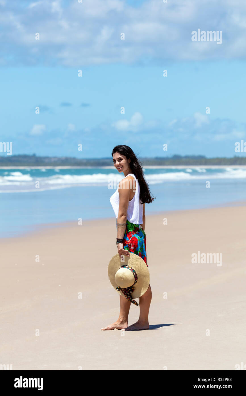 A beautiful young woman in a skirt, white top and straw hat on a white sand palm tree beach in the tropics Stock Photo