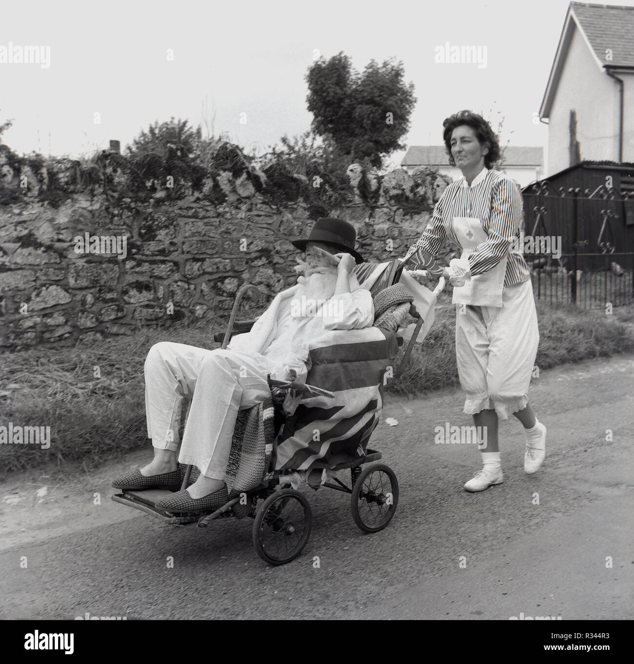 1967, pram race, Quainton, a lady dressed up as a nurse pushes a male competitor wearing hospital patient clothes, top hat and slippers, along a country road in the annual village pram race, Bucks, England, UK. Stock Photo