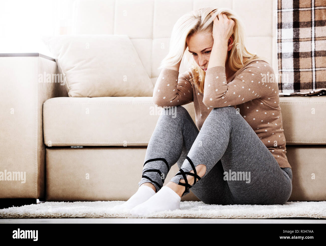 Depression. Middle aged barefoot woman sitting at the floor embracing her knees, near sofa at home, her head down, bored, troubled with domestic violence. Stock Photo