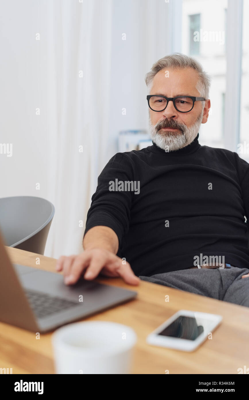 Businessman working at a laptop in the office sitting back relaxing in his chair as he browses online Stock Photo