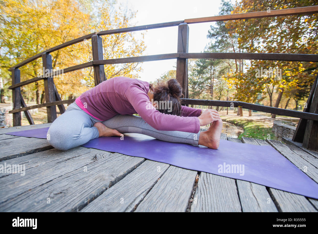 Young Sporty Woman Doing Yoga Exercise, Head To Knee Forward Bend Pose,  On Wooden Bridge At Park. Morning Autumn Day. Healthy Lifestyle. Stock Photo