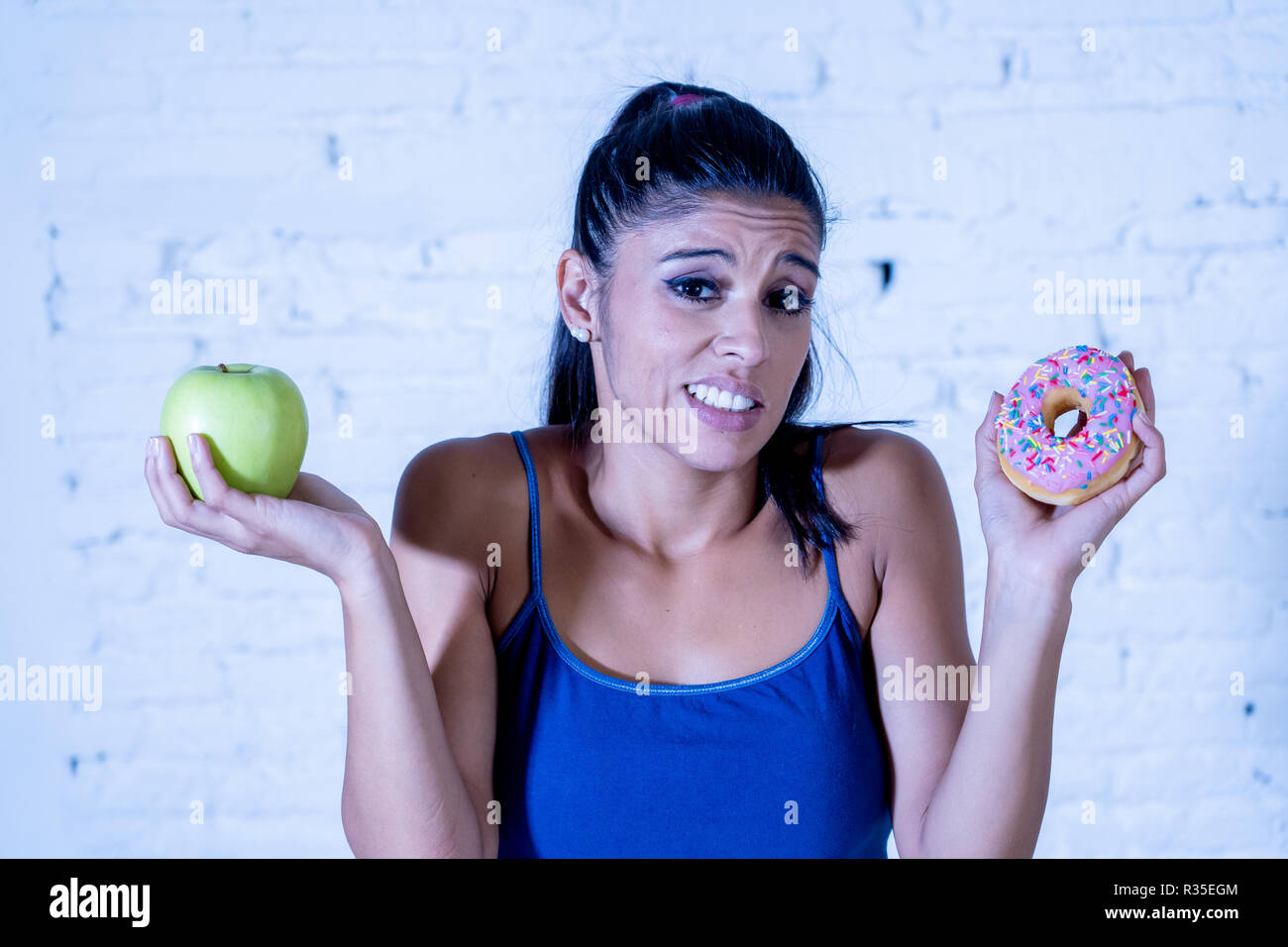 Beautiful young woman tempted having to make choice between apple and doughnut in healthy unhealthy food, detox eating, calories and diet concept. Stock Photo