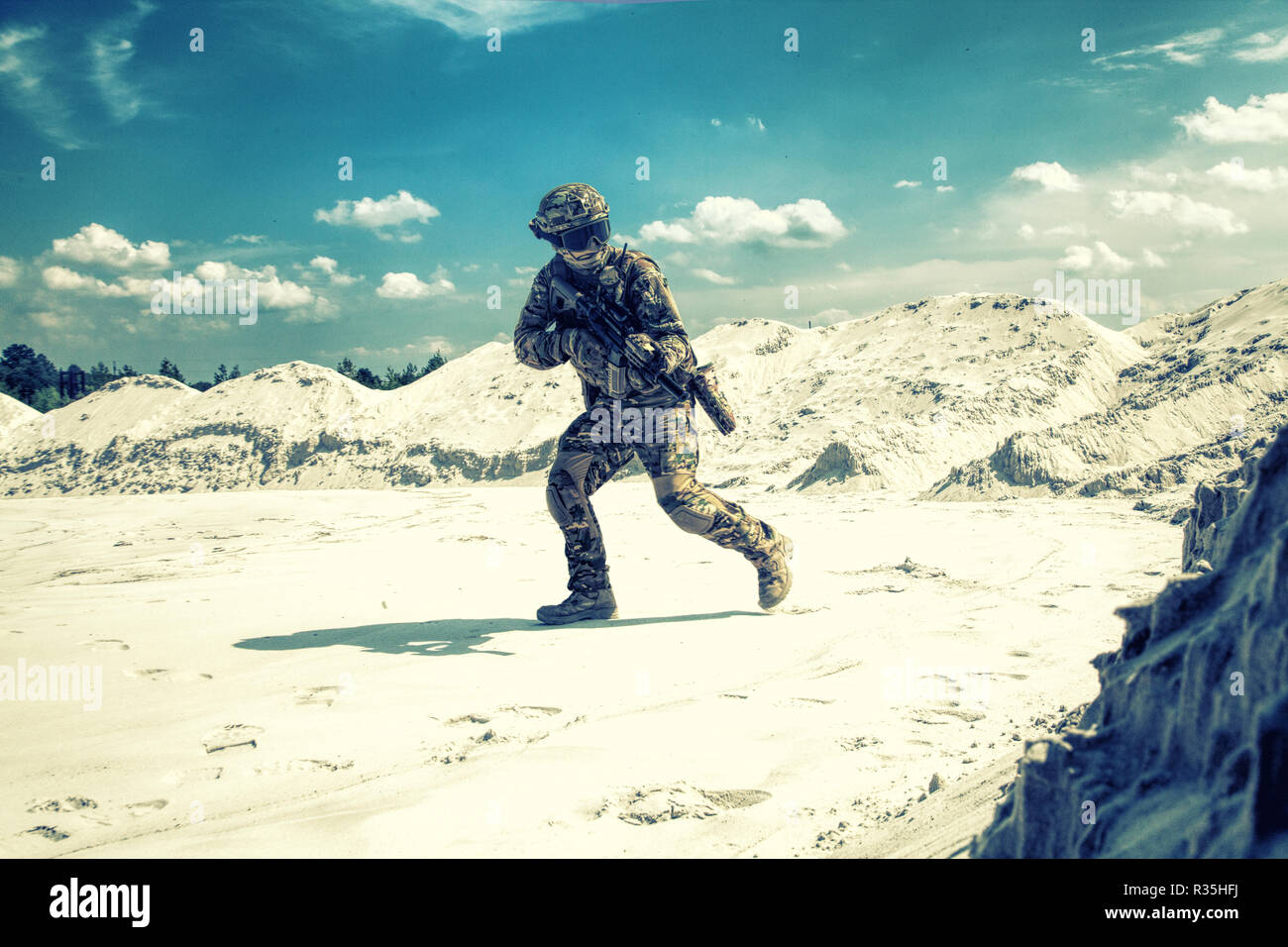 Man in military camouflage uniform with airsoft weapon bending down and running during armed conflict, military battle imitation in sandy or desert ar Stock Photo