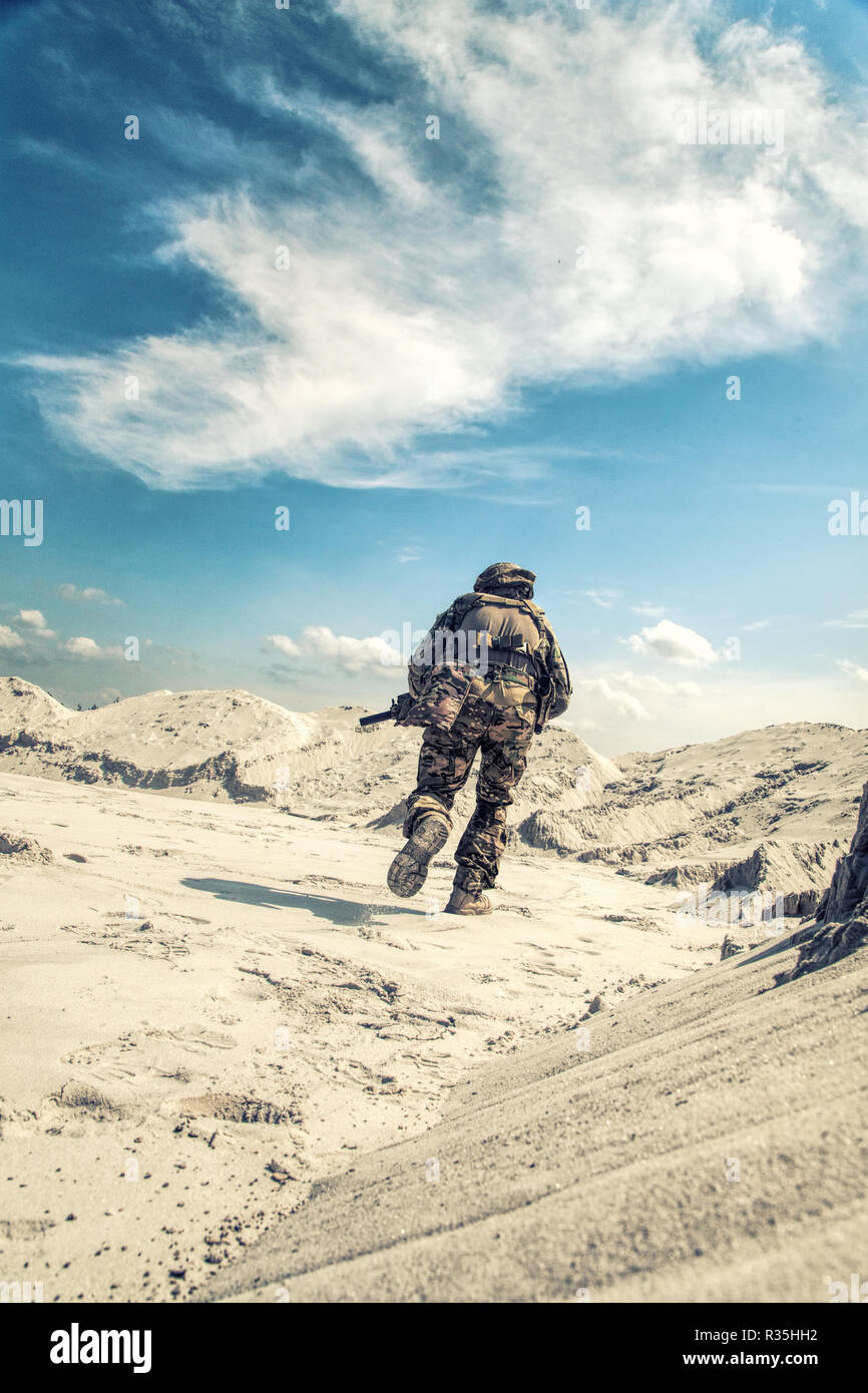 Man in military camouflage uniform with airsoft weapon bending down and running during armed conflict, military battle imitation in sandy or desert ar Stock Photo