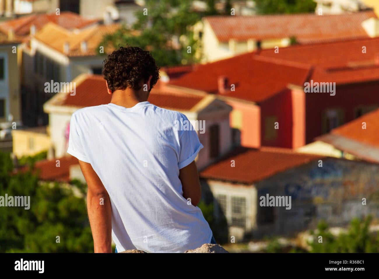 Athens, Greece - June 9, 2018: A young man sitting on a rock opposite the Acropolis of Athens is gazing towards the city buildings below Stock Photo