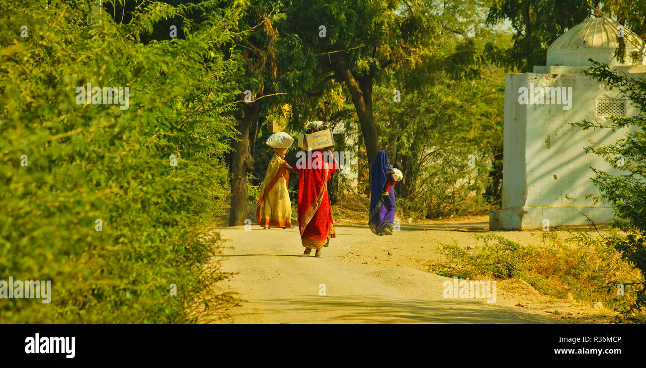 RAJASTHAN INDIA DUSTY ROAD AND THREE WOMEN WITH COLOURFUL SAREES AND LOADS ON THEIR HEADS Stock Photo