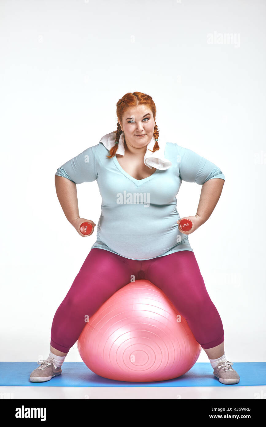 Red haired, chubby woman is sitting on the ball and keep the dumbbells Stock Photo