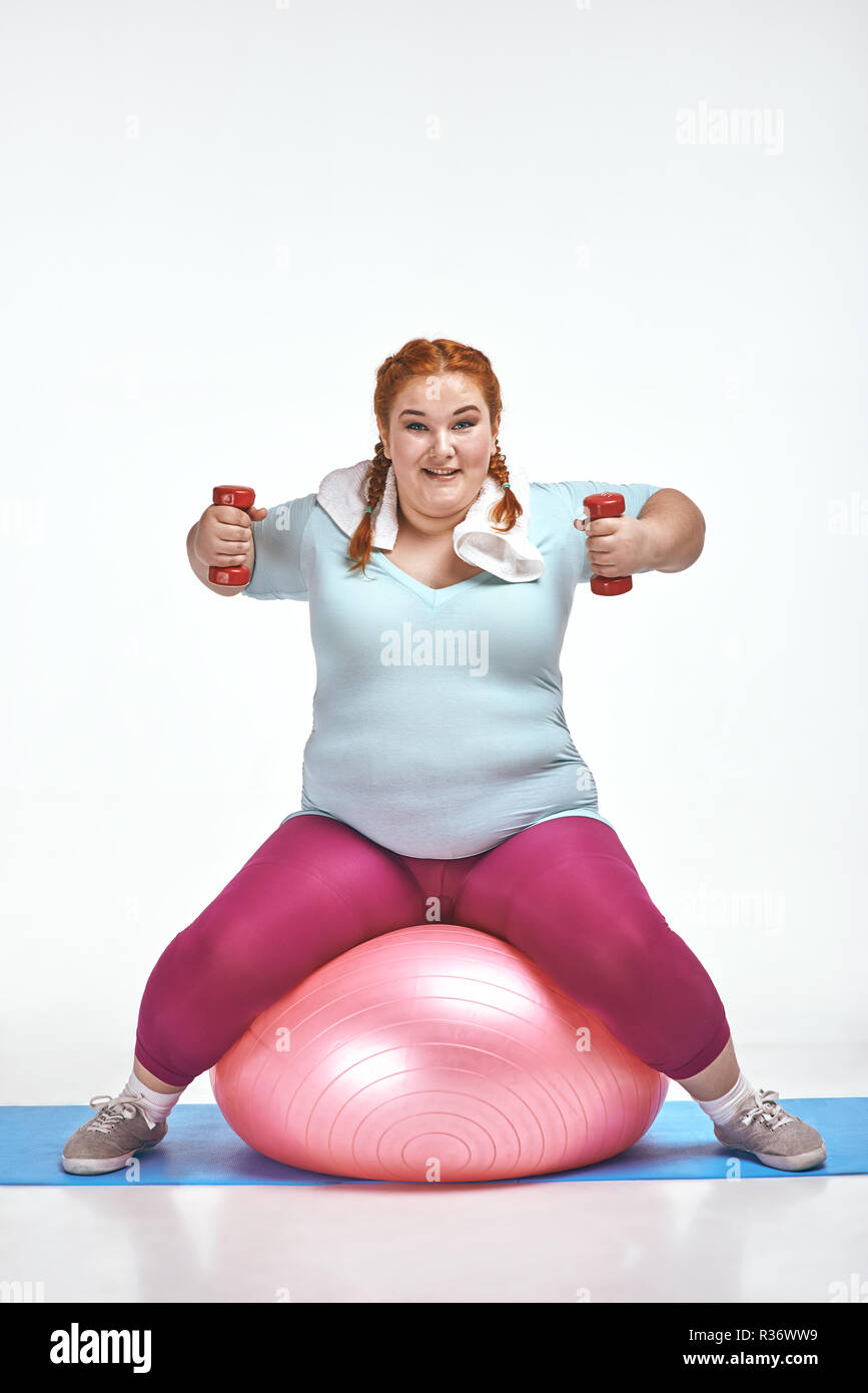 Red haired, chubby woman is sitting on the ball and keep the dumbbells Stock Photo