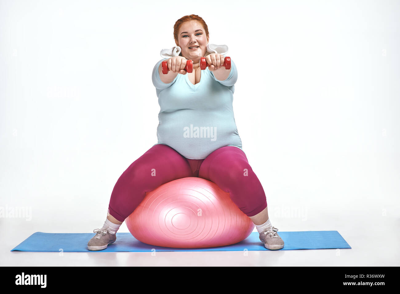 Red haired, chubby woman is sitting on the ball and keep the dumbbells Stock Photo