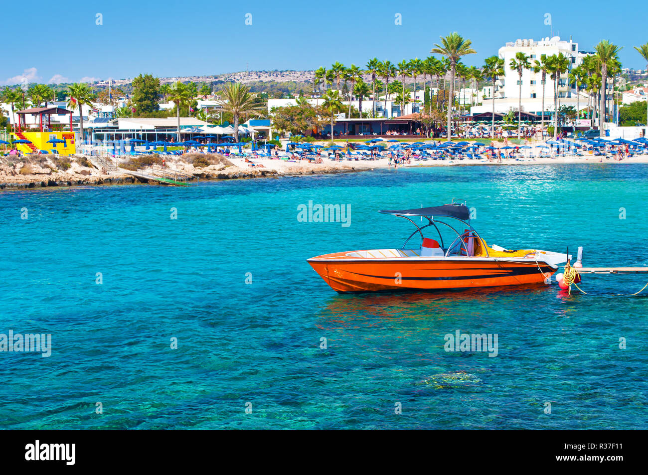 Image of one red speedboat near Vathia Gonia beach near Agia Napa, Cyprus. Sea with turquoise blue water, many green palm trees, houses on the backgro Stock Photo