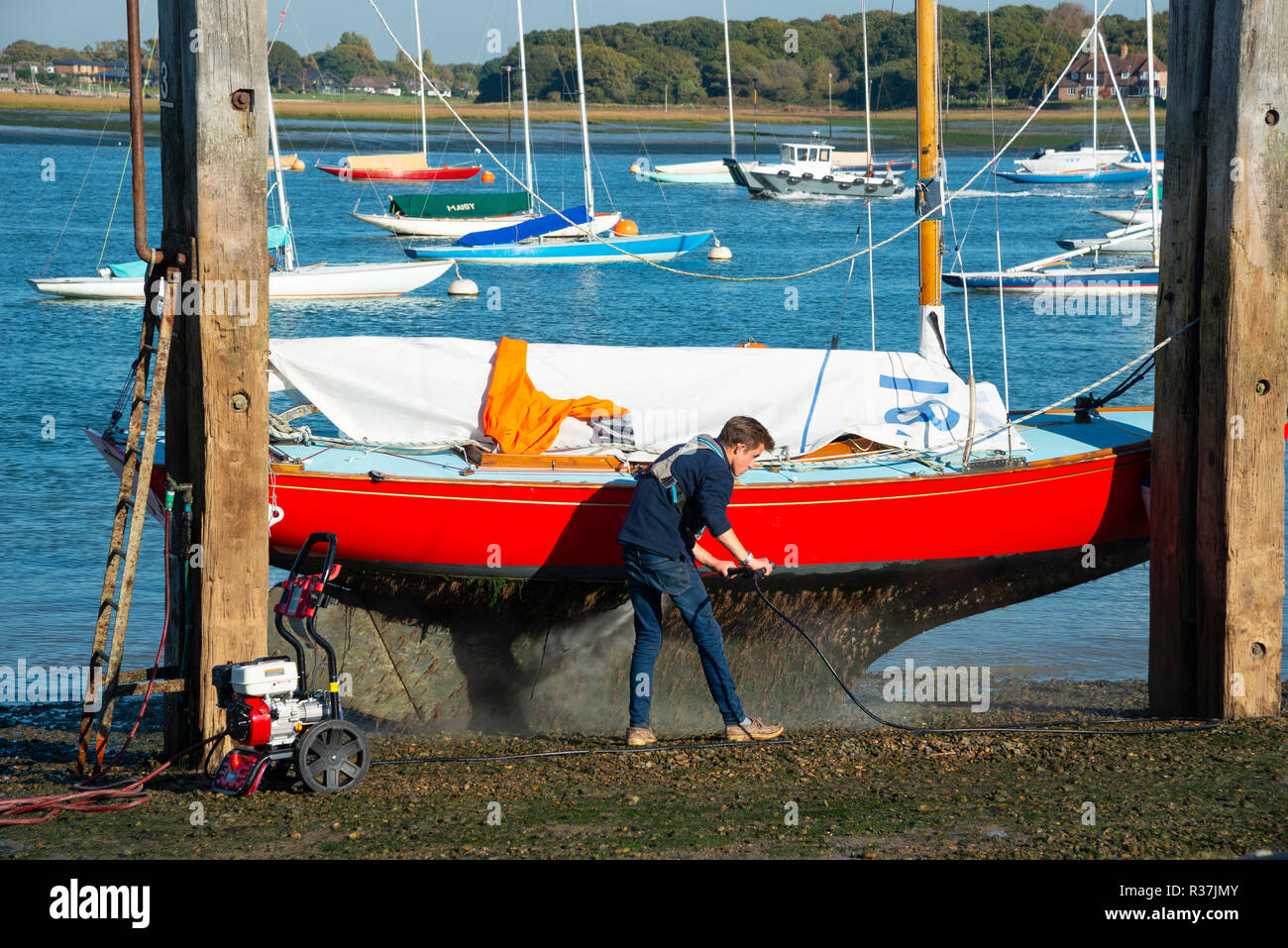 XOD Class yacht rests on dolphins while man jet washes hull on the hard at Itchenor, Chichester Harbour, West Sussex, England, UK Stock Photo