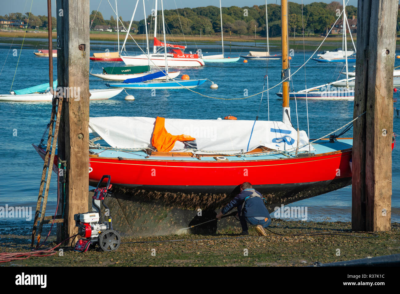 XOD Class yacht rests on dolphins while man jet washes hull on the hard at Itchenor, Chichester Harbour, West Sussex, England, UK Stock Photo