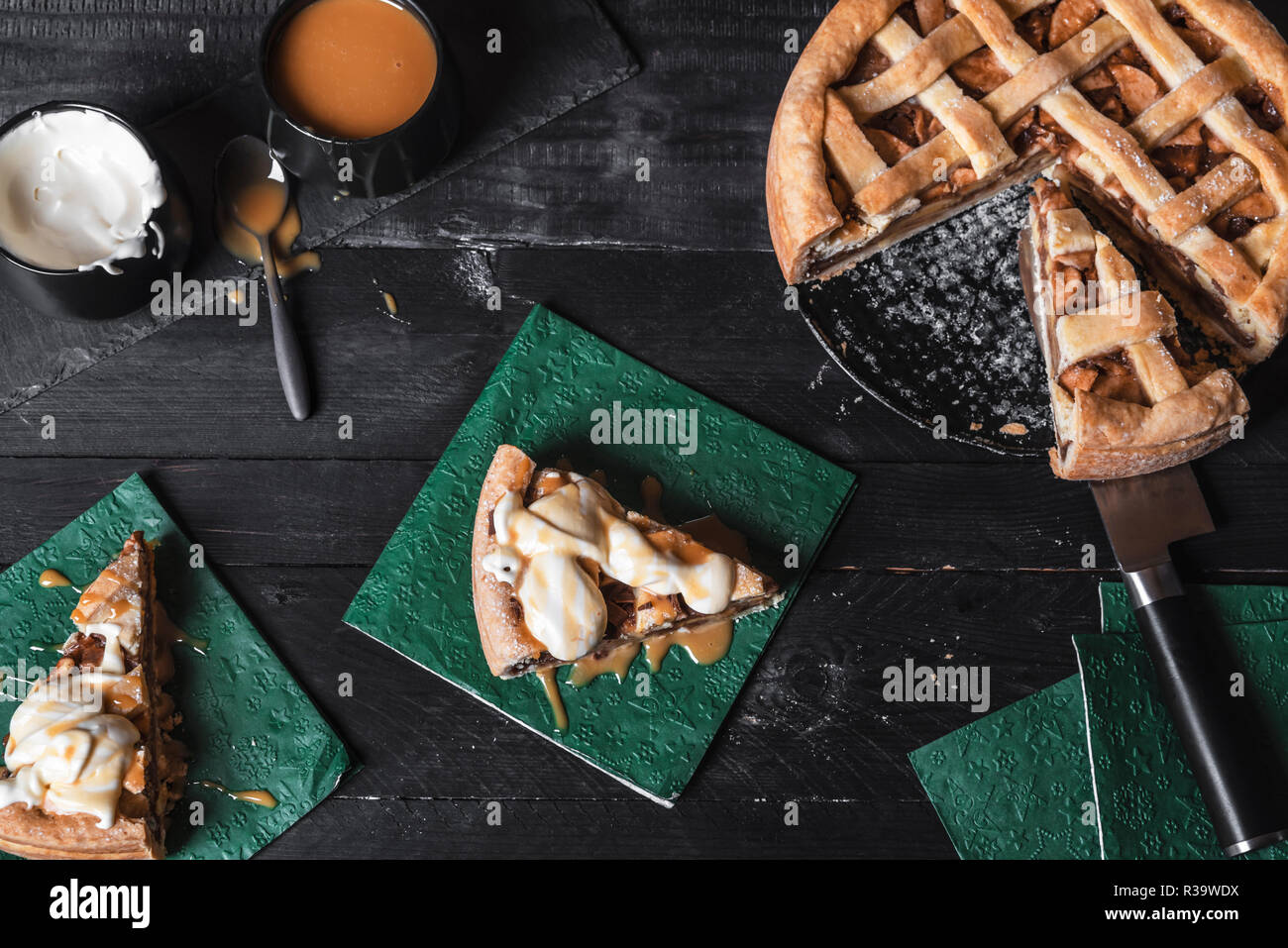 Above view of a rustic table with a sliced apple pie, and two pieces on napkins with cream and caramel sauce. Homemade cake. Traditional classic pie. Stock Photo