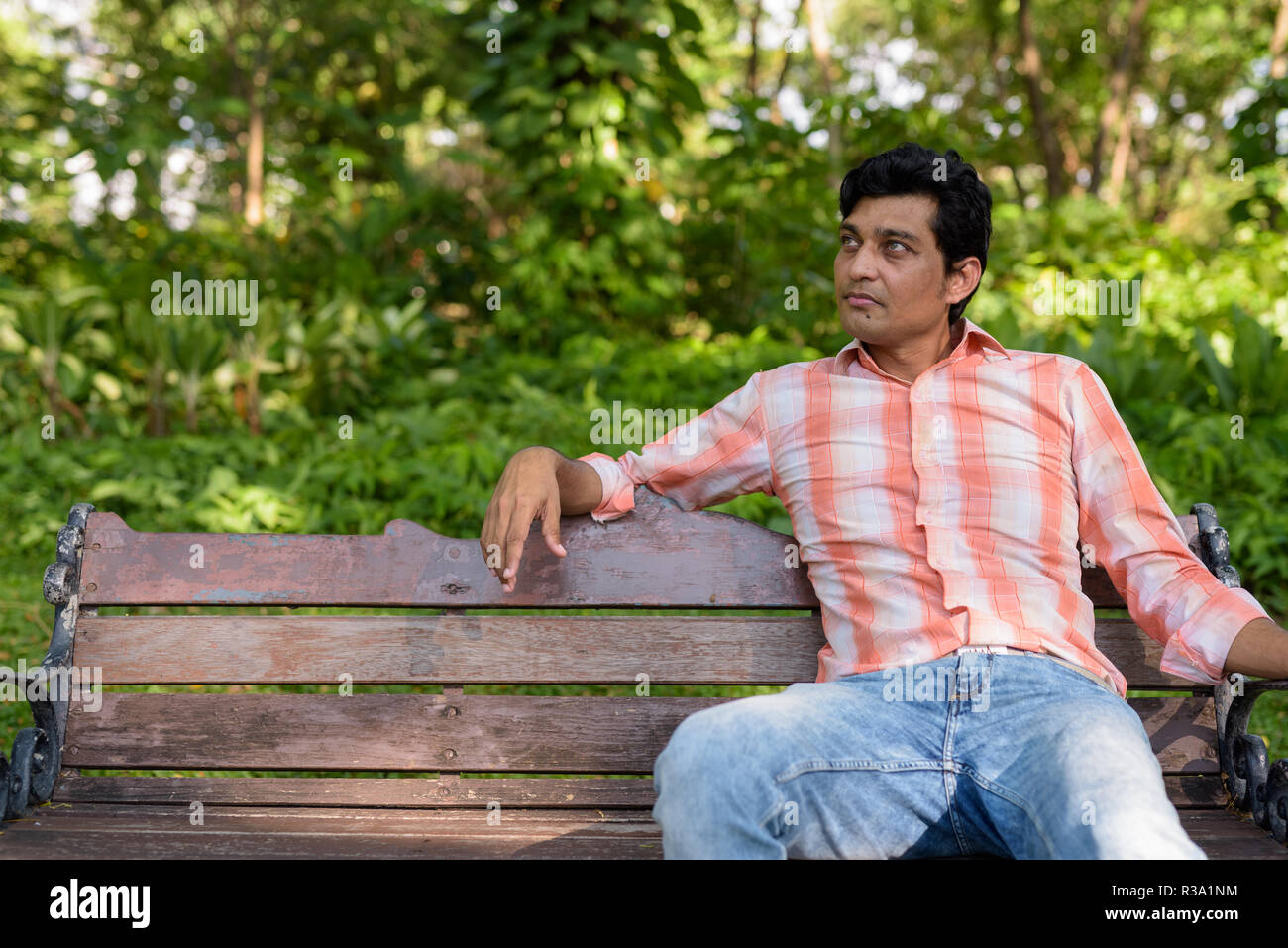 Indian man thinking while sitting on wooden bench in peaceful gr Stock Photo