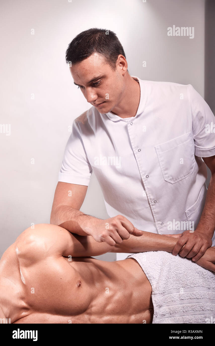 two young man, 20-29 years old, sports physiotherapy indoors in studio, photo shoot. Therapist masseur portrait massaging arm of muscular patient layi Stock Photo