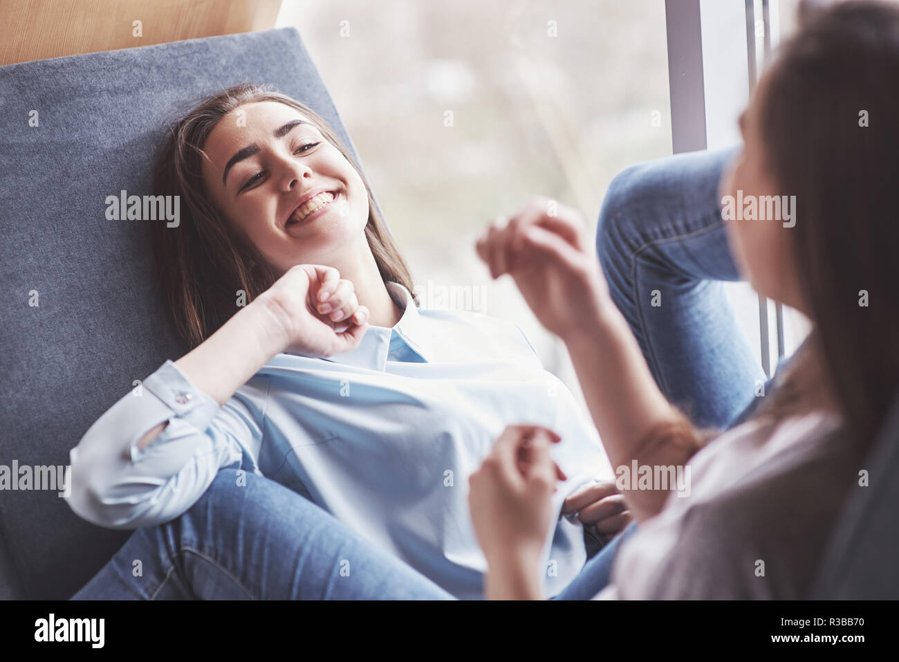Two twin girls sit in a tight armchair for them in the recreation area. Sisters posing and having fun spend time. Always join friendship and youth concept Stock Photo
