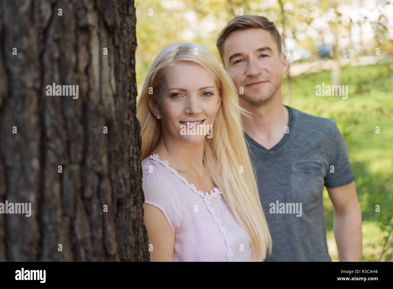 Attractive young couple outdoors in a park standing close together relaxing under a tree and smiling at the camera Stock Photo