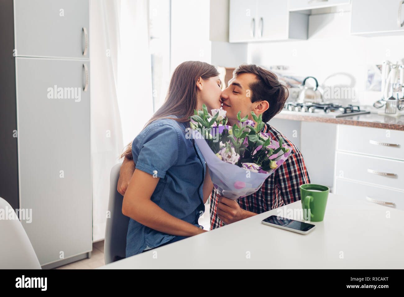 Young man gifting bouquet of flowers to his girlfriend in kitchen. Happy couple hugging. Romantic surprise at home Stock Photo