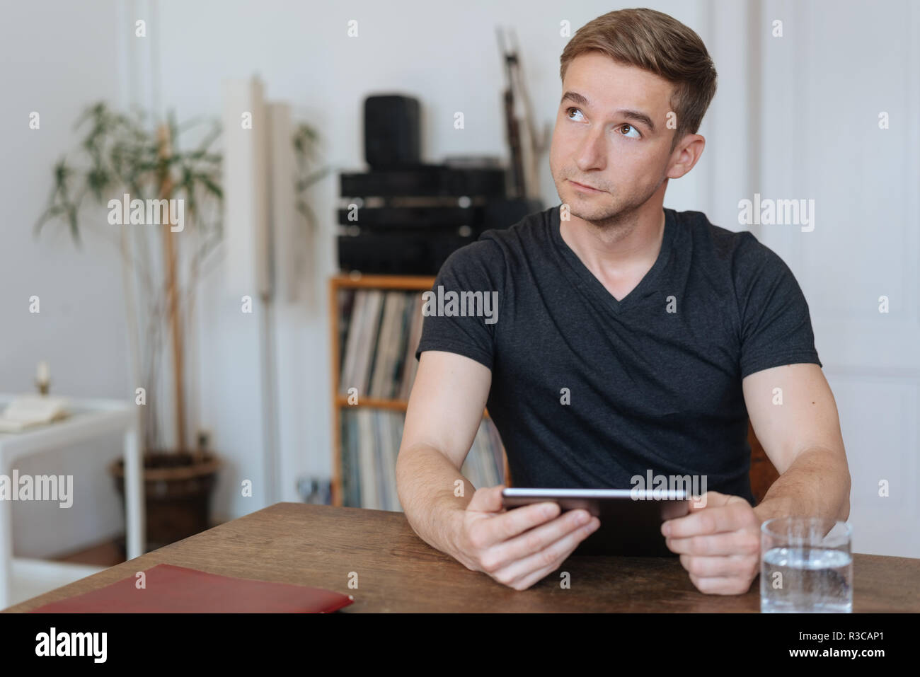 Young Man Sitting Deep In Thought At A Dining Table At Home Holding A Tablet Pc As He Looks Pensively To The Side Stock Photo Alamy